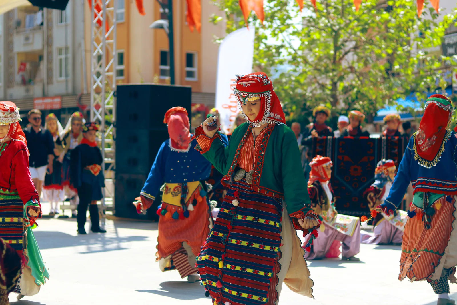 Dancers in striped, embroidered regional costume with red headscarves move in a line along a street at an outdoor festival, with a stage and onlookers behind. Not taken at this venue.
