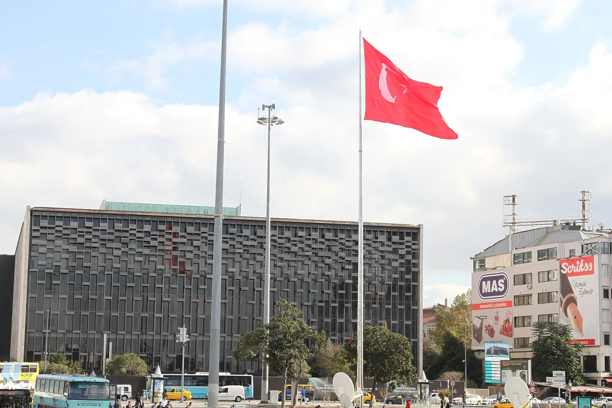 The dark, grid-patterned facade of the Atatürk Cultural Center beside tall flagpoles with a large Turkish flag