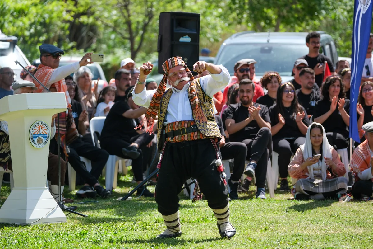 A man in a patterned vest, sash and baggy trousers dances in a park clearing in Antalya, with a seated audience clapping behind him