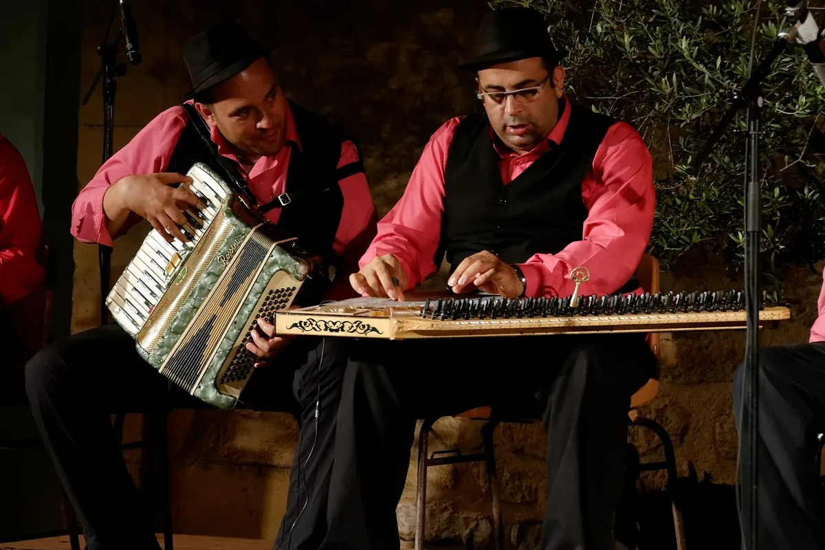 Two musicians in pink shirts and black waistcoats play a qanun and an accordion on a dimly lit stage