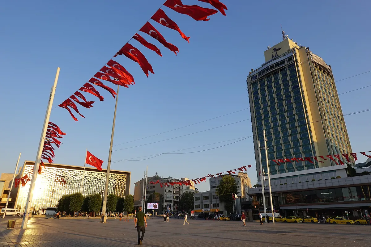Taksim Square on a sunny day, with a modern hotel tower rising behind strings of flags and people crossing the paved square
