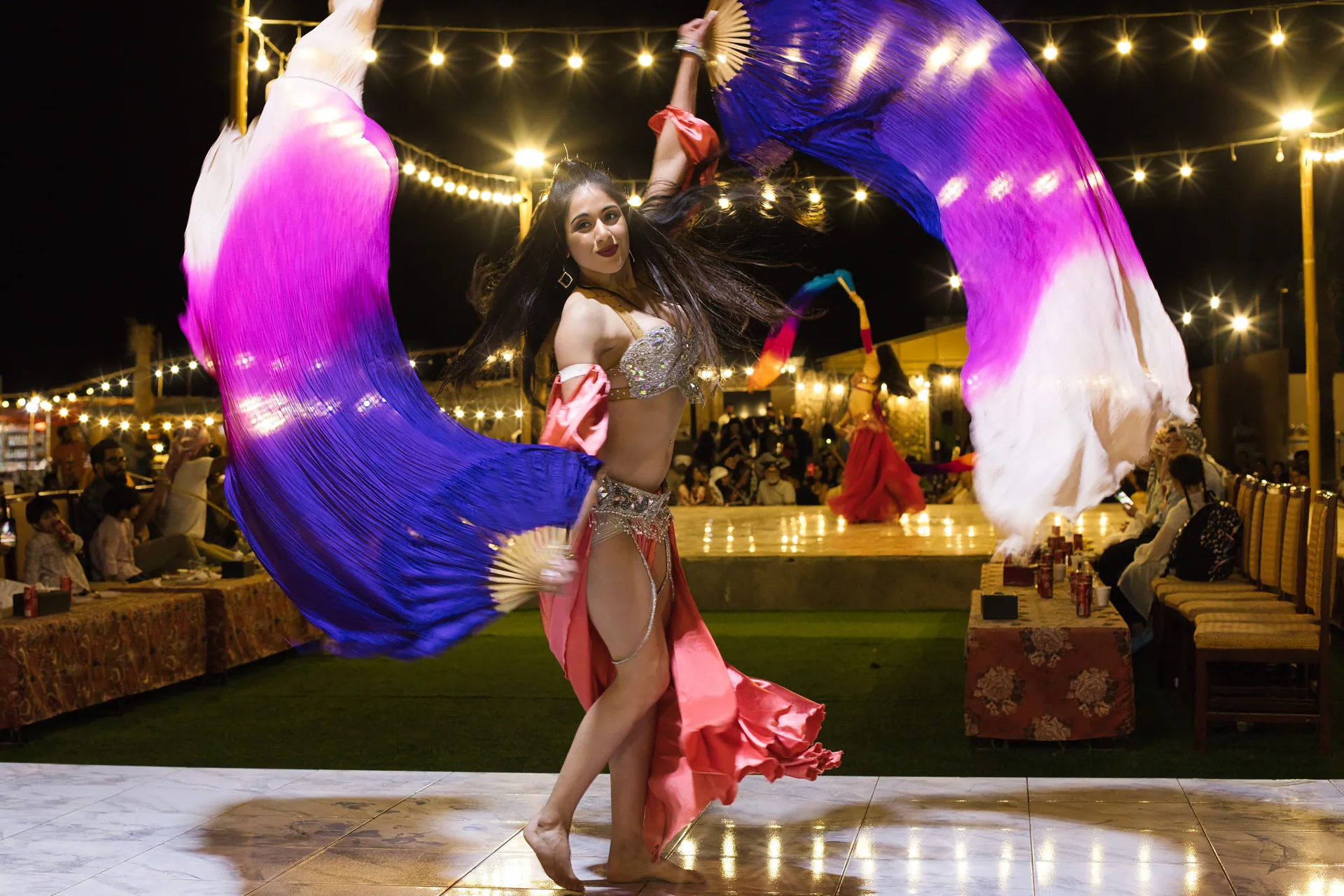 A belly dancer in a coral skirt and silver beaded top sweeps two large violet, magenta and white silk fans around her on a lit stage at night, with strings of lights above and diners at tables behind. A general belly dance photograph, not taken at this venue.