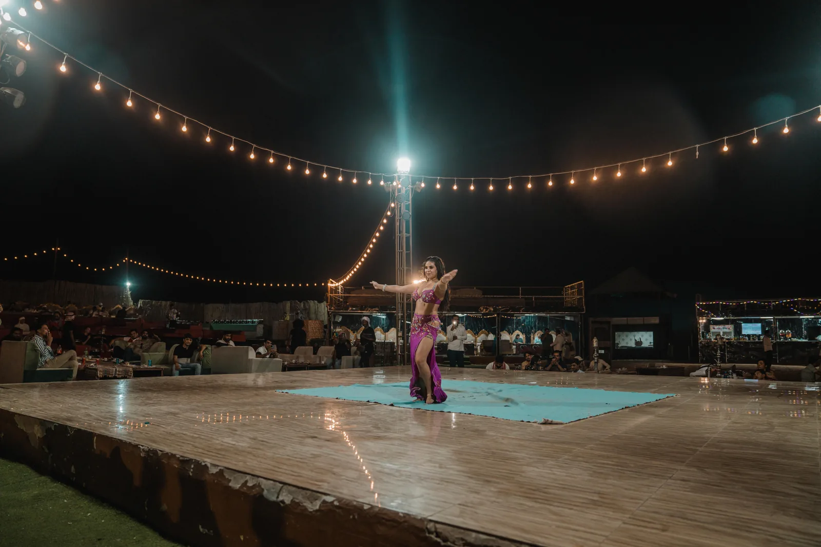 A belly dancer in a magenta costume on a small turquoise rug at the centre of a wide wooden stage, under arcs of string lights, with seated diners around the edge. A general photograph, not taken at this venue.