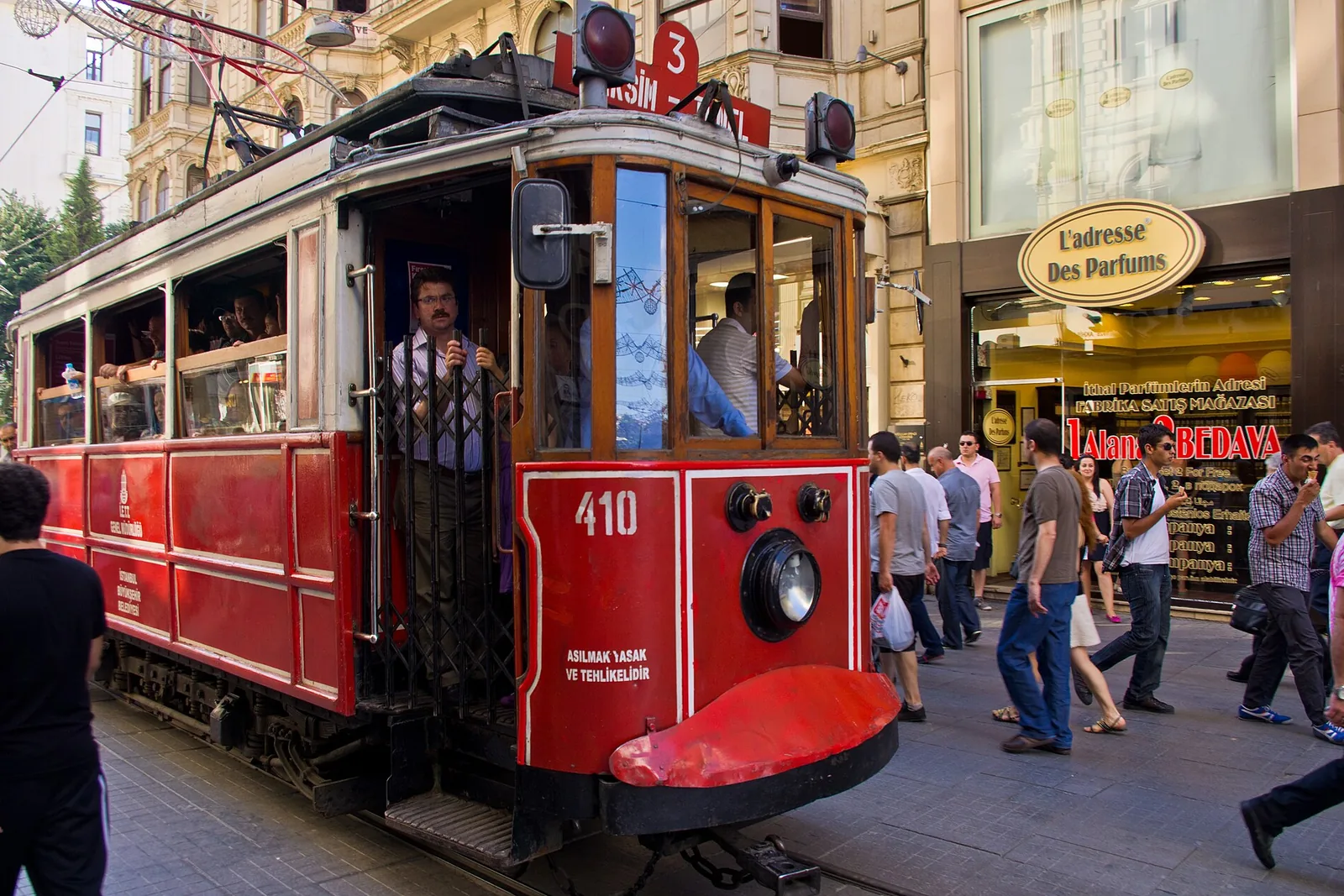 A red historic tram, number 410, with its driver at the open door, on a crowded pedestrian street lined with old stone shopfronts on İstiklal Avenue.
